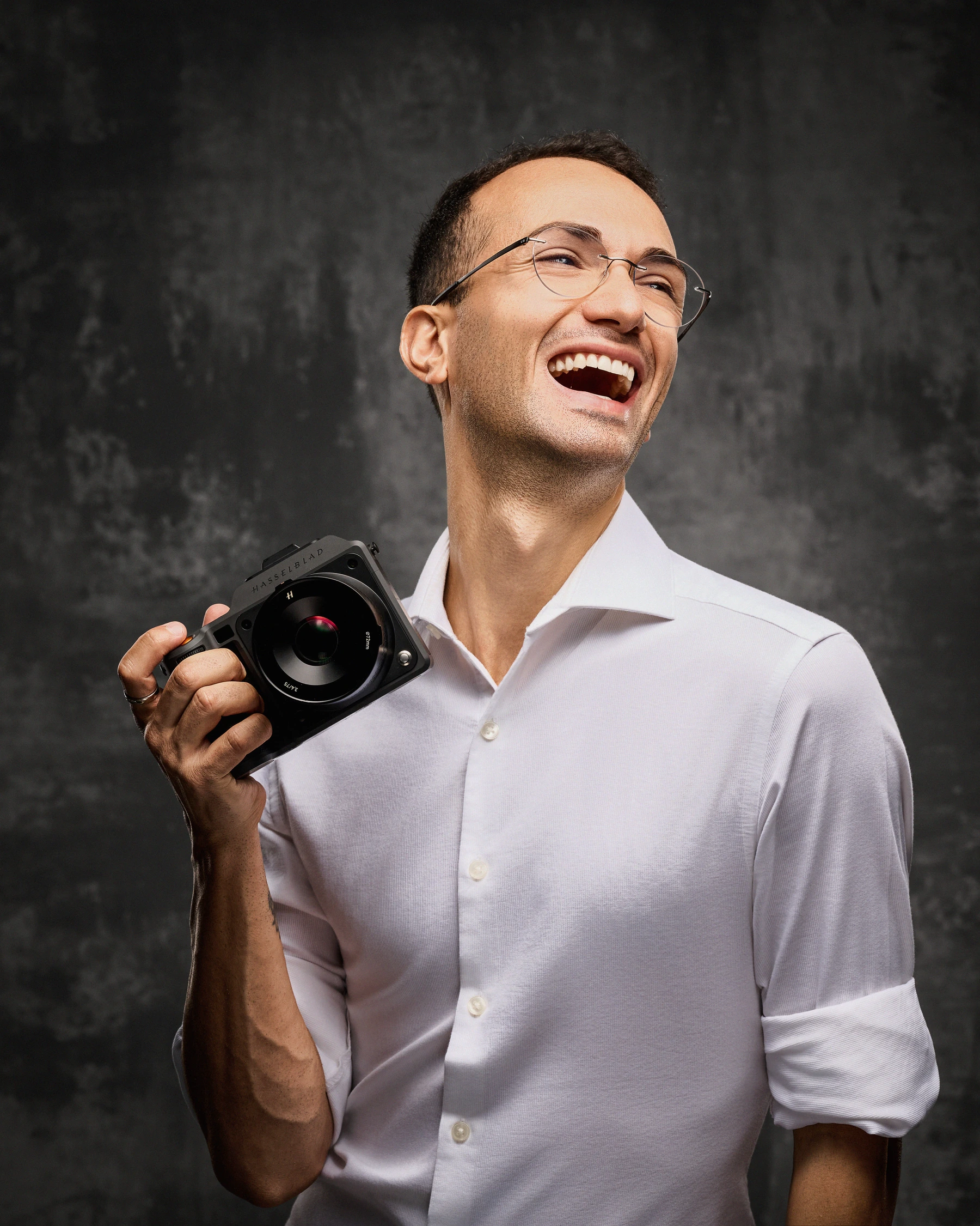 Luca laughing while holding his camera, in a white shirt against a textured gray backdrop