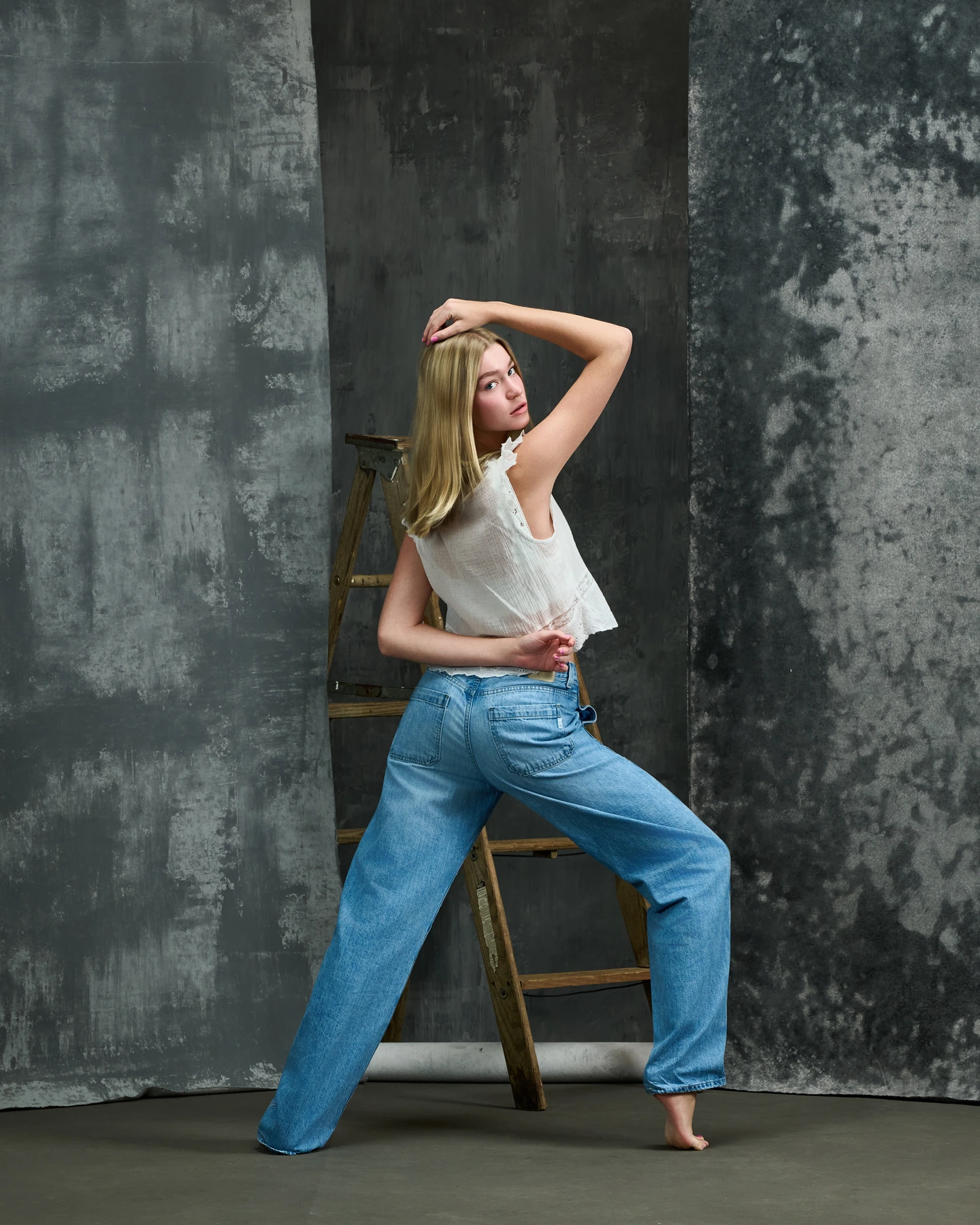 Dancer in jeans and a white blouse beside a ladder and textured gray backdrops