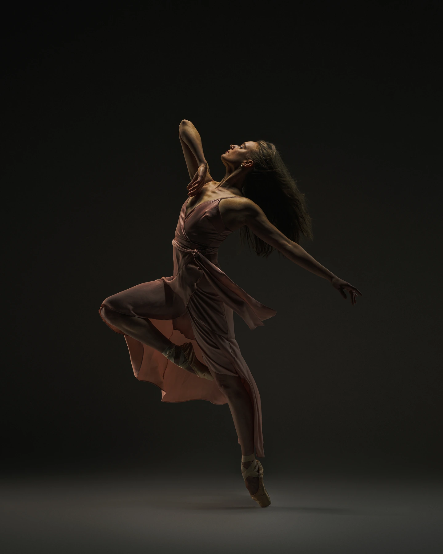Ballerina in a dusty rose dress illuminated against a dark backdrop