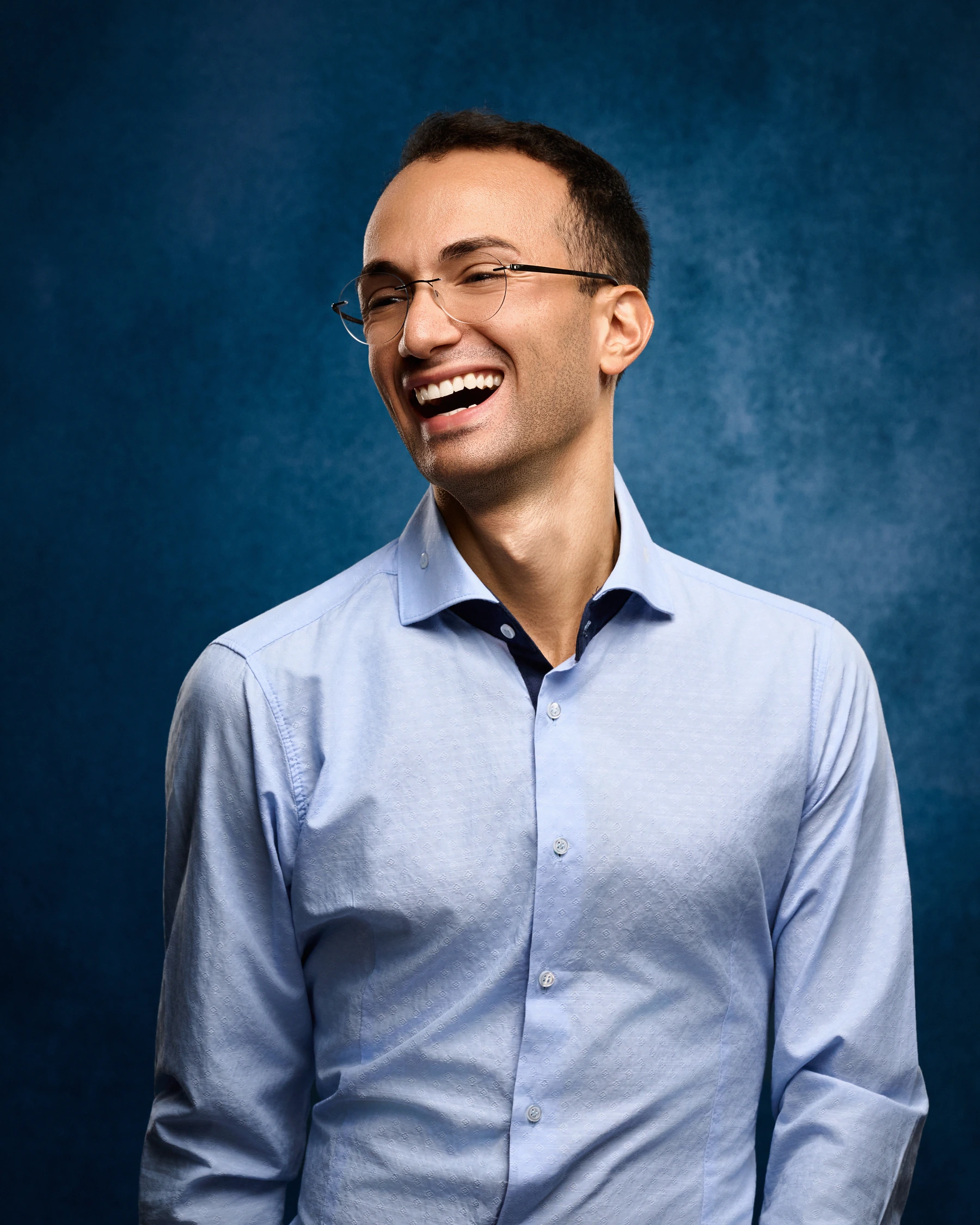 Luca laughing in a light blue shirt against a textured blue backdrop