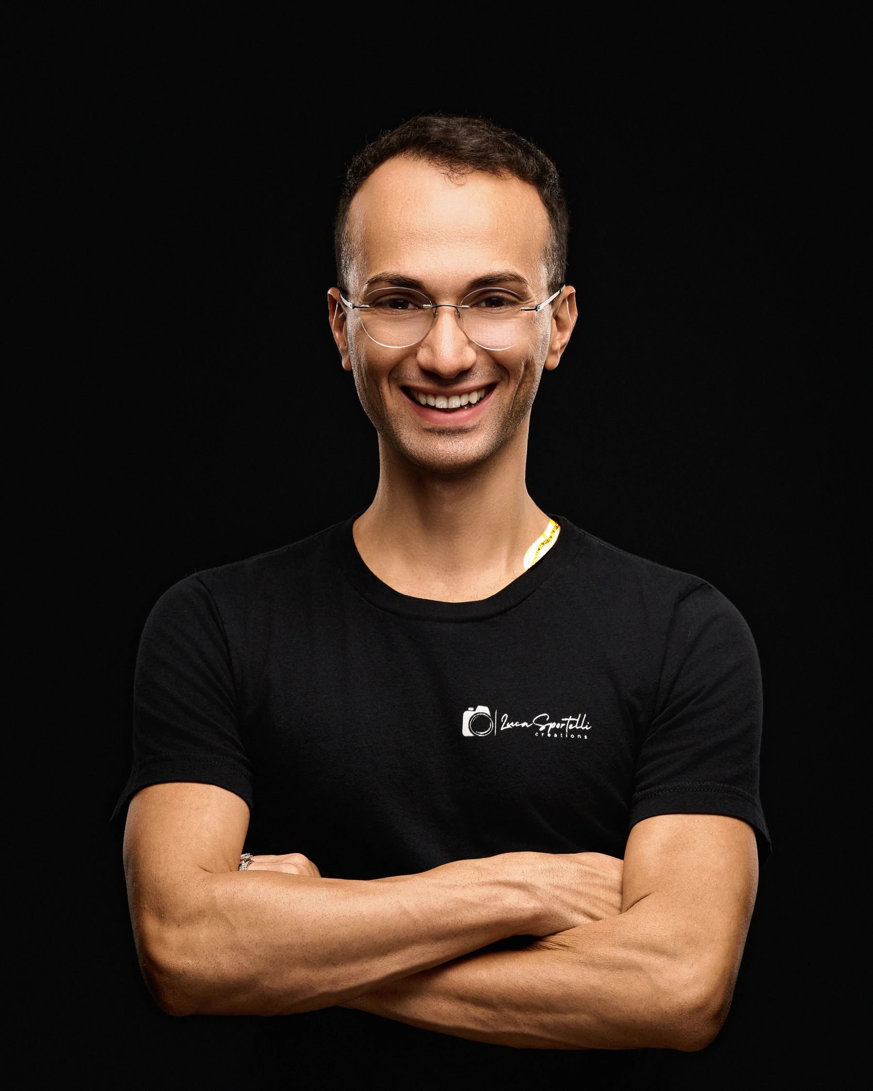 Luca smiling with his arms crossed, wearing his studio logo shirt against black