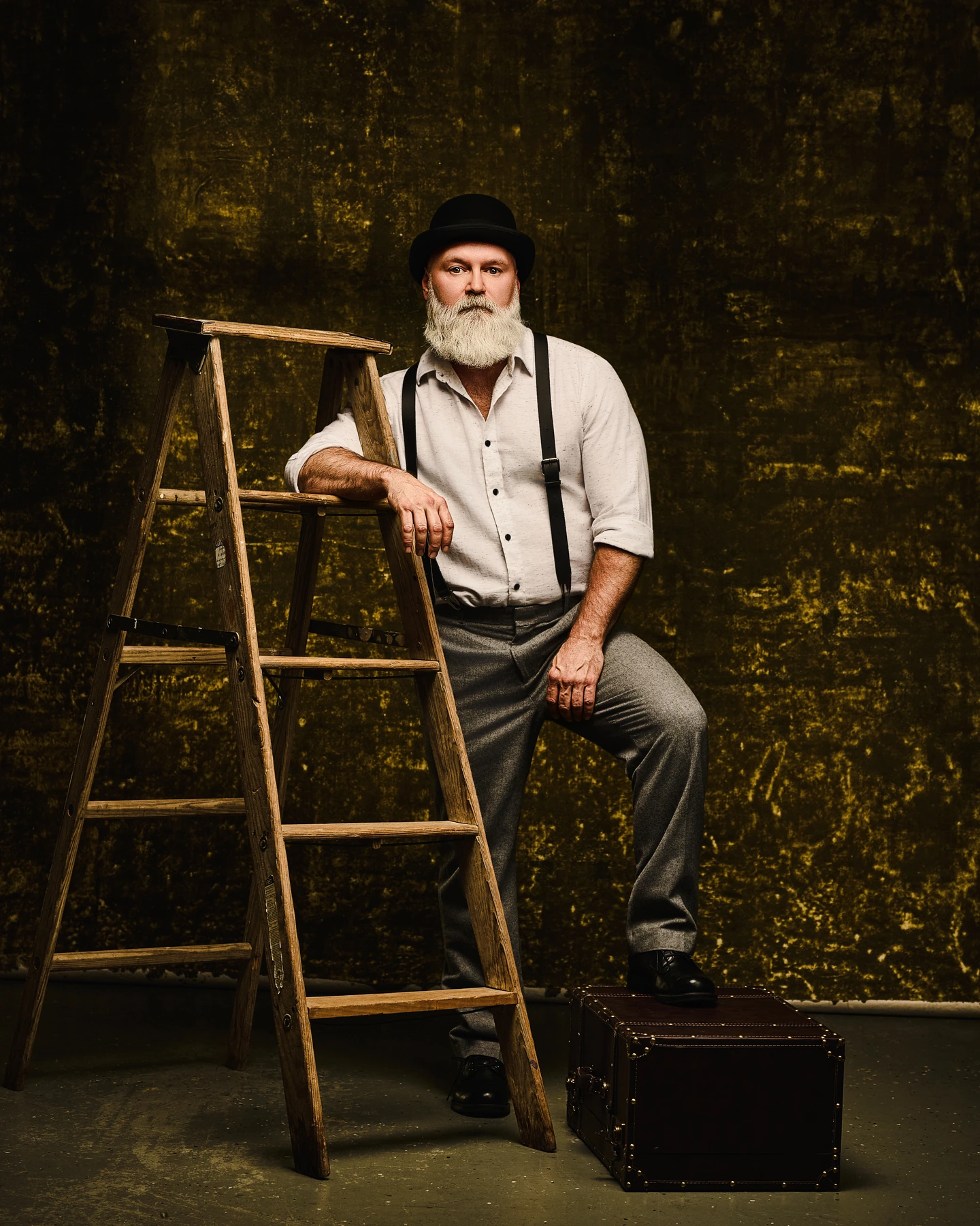 Bearded man in a black hat beside a wooden ladder against a textured olive backdrop