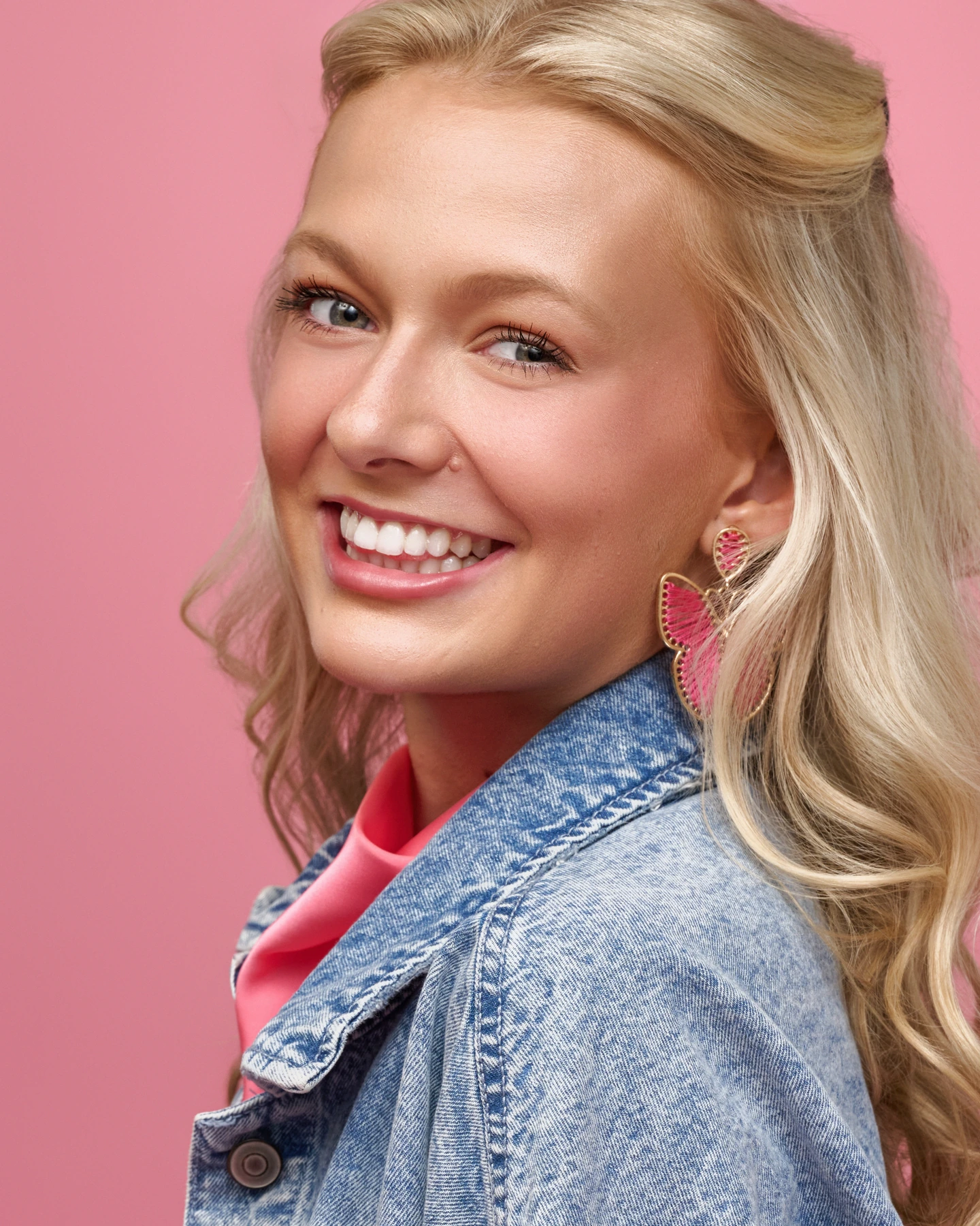 Smiling portrait with butterfly earrings and a denim jacket against pink