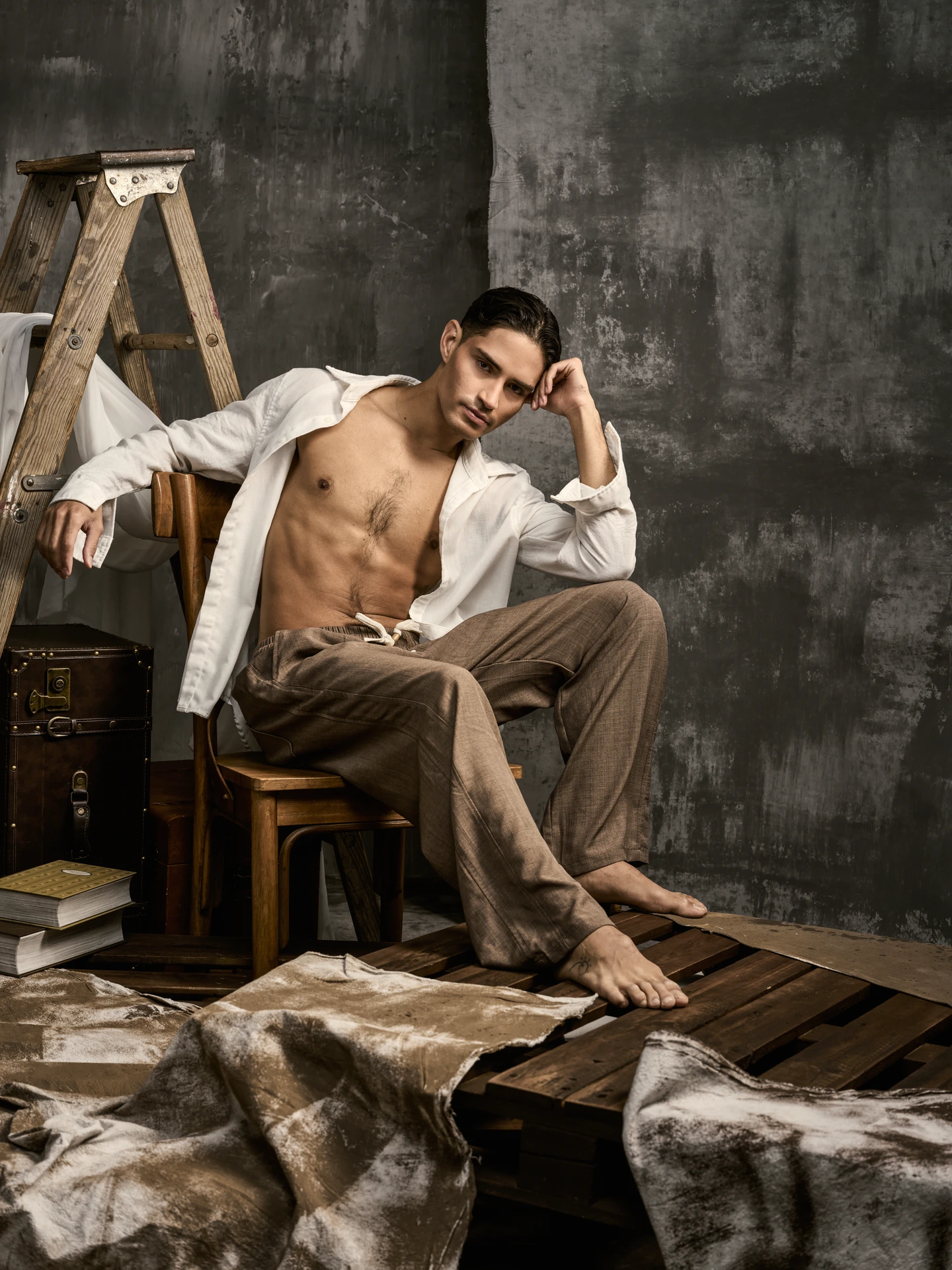 Seated portrait in an open white shirt and brown trousers among vintage studio props