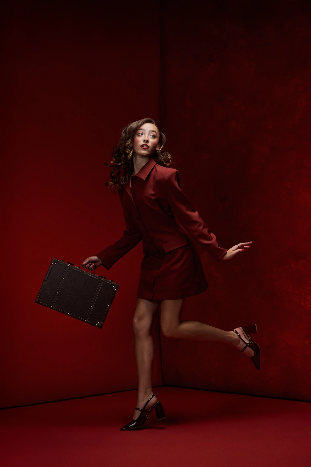 Fashion portrait in a red skirt suit holding a suitcase against deep red